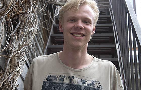 Josh standing in front of staircase covered on one side with vines. smiling, spiked blond hair