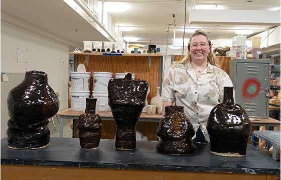 Danielle standing in white sweater behind table stacked with her pottery