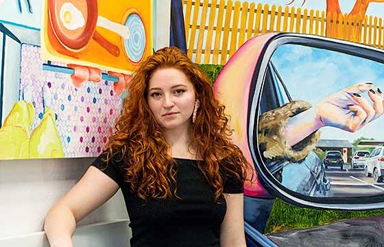 Chloe in black shirt and red hair down stands in front of paintings